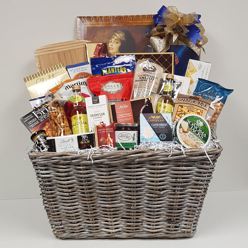 A large basket filled with chocolates, biscuits, pretzels, apple chips, crackers and cheeses, baked brie topping, smoked salmon, large-sized cutting board, flavoured sparkling water, red pepper jelly, mustard, snack mix, nuts, savoury shortbread, salami and more.