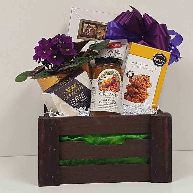 A wooden crate with chocolate, crackers and cheese, shortbread, gourmet chutney and a potted plant.