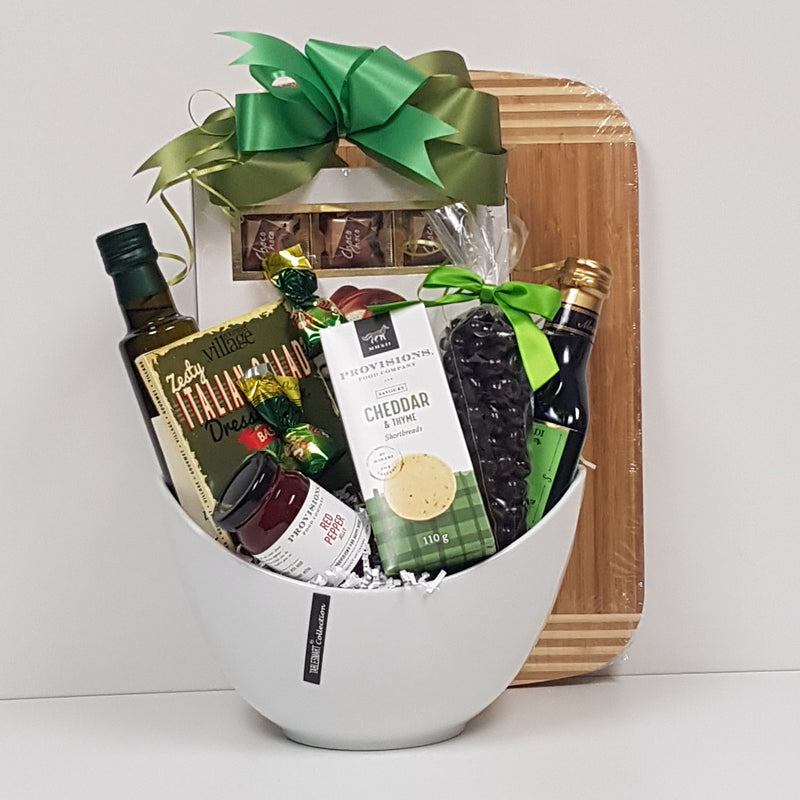 A ceramic bowl filled with gourmet oil and vinegar, savoury shortbread, red pepper jelly, dressing or dip mix, a cutting board and chocolate treats.
