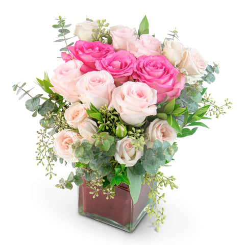 Glass cube arrangement of light and bright pink roses and spray roses with lush greens.